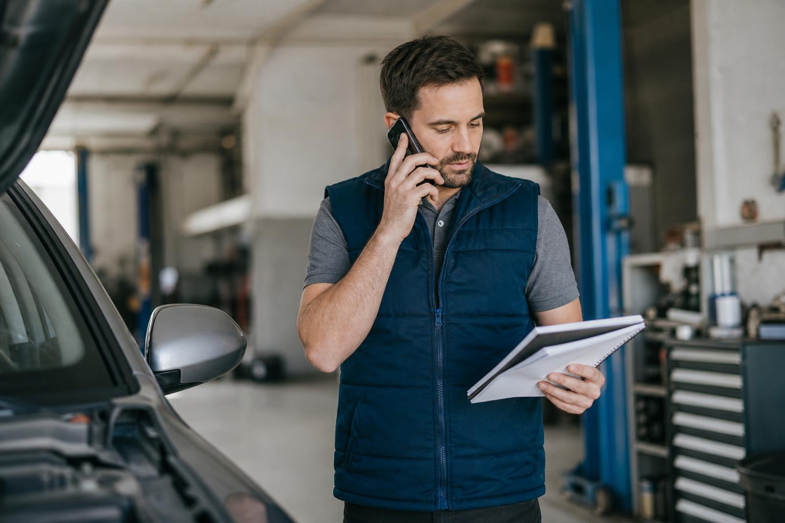 Automotive service manager taking a customer call in a workshop while checking job notes, the kind of call an automotive AI voice agent can handle